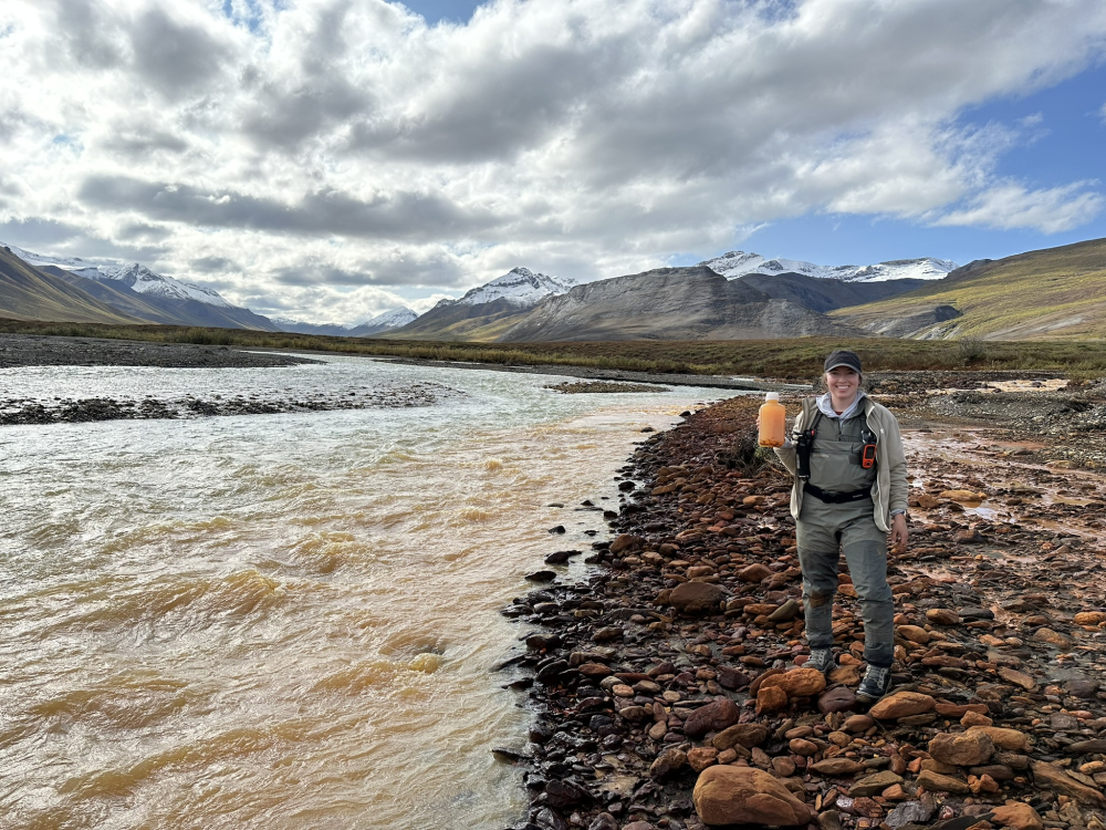 a woman holds up a bottle of very orange water next to a river that looks dirty orange, it is a very beautiful landscape